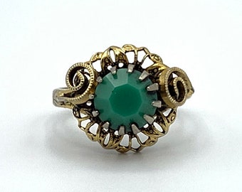 May include: Vintage ring featuring a large, faceted emerald-green stone set in a filigree gold-tone metal setting. The ring has intricate scrollwork details on the sides, creating an elegant, antique aesthetic.