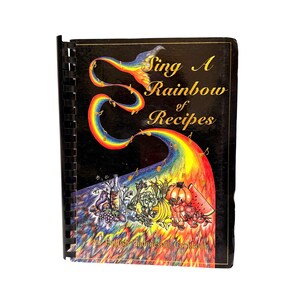 Könnte beinhalten: Ein spiralgebundenes Kochbuch mit dem Titel "Sing A Rainbow of Recipes" mit schwarzem Einband. Das Cover zeigt einen Regenbogen, Noten und Illustrationen von Lebensmitteln. Der Text "An English/Indonesian Cookbook" befindet sich unten.