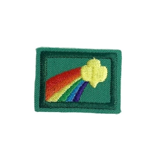 Pode incluir: Remendo bordado com um arco-íris que se estende da esquerda para a direita, terminando em uma forma de trevo amarelo. O arco-íris inclui vermelho, laranja, amarelo, verde, azul e roxo. O remendo é retangular com uma borda e fundo verdes.