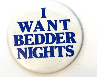 May include: A white button with the blue text "I WANT BEDDER NIGHTS". The button is round and has a slightly worn appearance. The text is centered and easy to read.