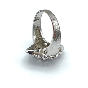 May include: A silver-colored ring with a detailed, textured design. The ring features a central, ornate setting with a cluster of small, dark stones. The band has a smooth, polished finish. The ring is shown against a white background.