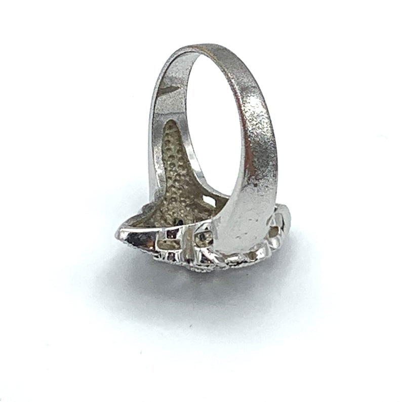 May include: A silver-colored ring with a detailed, textured design. The ring features a central, ornate setting with a cluster of small, dark stones. The band has a smooth, polished finish. The ring is shown against a white background.