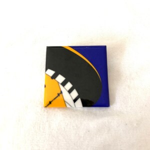May include: Square ceramic tile with a graphic design featuring a black, yellow, and white circular shape against a blue background. The design is reminiscent of a stylized wheel or clock face.