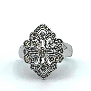 May include: A silver-toned ring with an ornate, symmetrical design. The ring's centerpiece features a detailed pattern of small, dark stones arranged in a floral or cross-like shape. The band is smooth and simple.