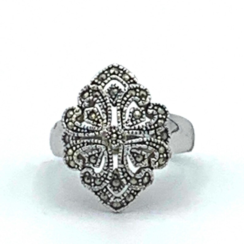May include: A silver-toned ring with an ornate, symmetrical design. The ring's centerpiece features a detailed pattern of small, dark stones arranged in a floral or cross-like shape. The band is smooth and simple.