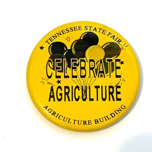 May include: A round, yellow button with the text "Tennessee State Fair" at the top. The button features a graphic of balloons and the words "Celebrate Agriculture" in black. The text "Agriculture Building" is at the bottom.