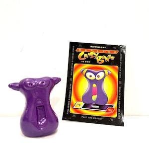 May include: A purple Crazy Bones figure with a unique shape, next to a collector sticker card. The card features a cartoon monster with the text "WOW" and "#8". The McDonald's Monster Crazy Bones collectible is on a white background.