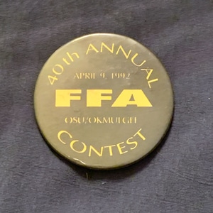 May include: A round, olive-green button with gold lettering. The text reads "40th ANNUAL" at the top, "APRIL 9, 1992" in the center, and "FFA" in bold yellow letters. Below, it says "OSU/OKMULGEE CONTEST".