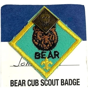 May include: A Bear Cub Scout badge with a brown bear design on a teal background, framed by a yellow border. A smaller metal badge with a bear face is on top. The text "BEAR" and "BEAR CUB SCOUT BADGE" are visible.
