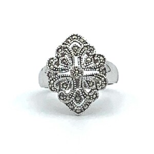 May include: A silver ring with a detailed, ornate design. The ring's centerpiece is a symmetrical, floral-like pattern, encrusted with small, dark stones. The band is smooth and polished, creating a contrast with the textured design. The ring is set against a white background.