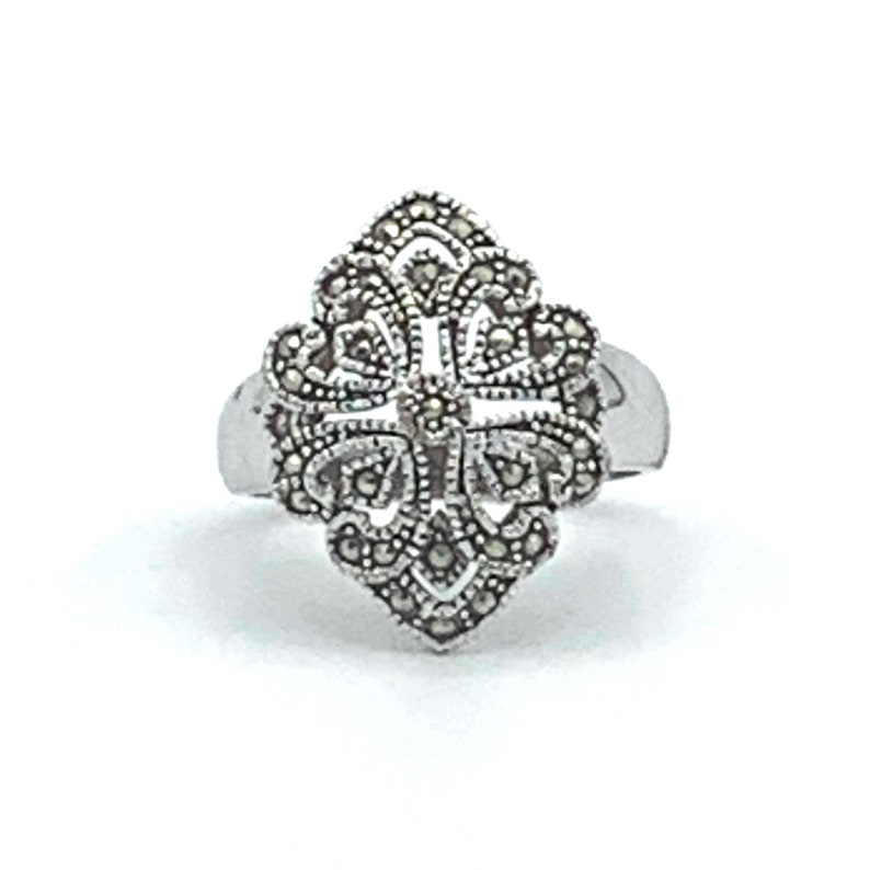 May include: A silver ring with a detailed, ornate design. The ring's centerpiece is a symmetrical, floral-like pattern, encrusted with small, dark stones. The band is smooth and polished, creating a contrast with the textured design. The ring is set against a white background.