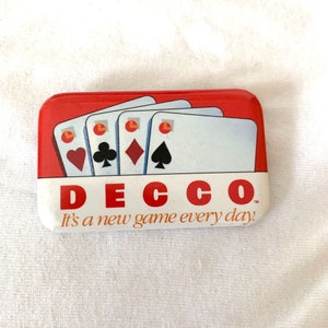 Puede incluir: Un pin rectangular con la parte superior roja e inferior blanca. La parte superior presenta cuatro cartas de juego con palos: corazones, tréboles, diamantes y picas. El texto "DECCO" está en rojo, y debajo, "It's a new game every day!"