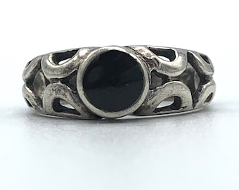 May include: A silver-toned ring with an openwork design and a central, circular black stone. The ring's band features a repeating pattern of curved shapes, creating an intricate and decorative look. The black stone provides a striking contrast to the silver metal.