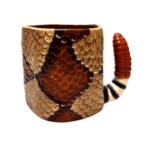 Könnte beinhalten: Ein Keramikbecher mit einer strukturierten Schlangenschuppen-Optik in Beige- und Dunkelbrauntönen. Der Henkel ist wie eine Klapperschlangenschwanz geformt, in Rotbraun gehalten und mit schwarz-weißen Streifen an der Basis versehen.
