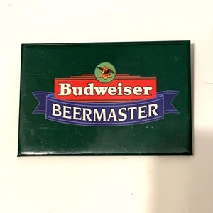 Puede incluir: Un imán rectangular verde Budweiser BeerMaster. El imán presenta el logotipo de Budweiser con un fondo rojo y texto blanco, y una pancarta azul con el texto blanco "BEERMASTER".