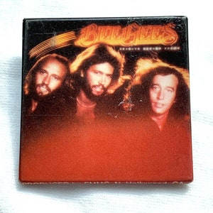 Puede incluir: Pin cuadrado con los Bee Gees. El pin tiene un borde negro y un fondo degradado rojo y naranja. Los miembros de la banda están representados en el centro. El texto "Bee Gees" está en la parte superior.