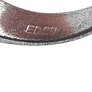 May include: Close-up of a silver-colored metal ring with the engraved text "EDCO". The ring has a textured surface and a slightly curved shape, with the text appearing in a sans-serif font.