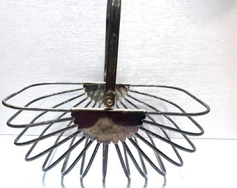 May include: A silver-toned metal basket with a handle. The basket has a fan-like design with curved wires radiating from the base. The handle is arched and attached to the basket with decorative floral-shaped accents. The basket is empty.