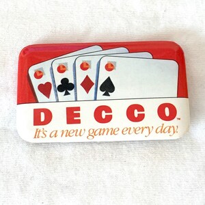 Puede incluir: Un pin rectangular con fondo rojo y sección inferior blanca. El pin presenta cuatro naipes y la palabra "DECCO" en letras rojas. El texto "It's a new game every day" está debajo de la palabra DECCO.