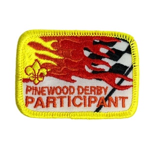 Może przedstawiać: Haftowana naszywka z żółtą obwódką i napisem "Pinewood Derby Participant". Naszywka przedstawia czerwono-żółty płomień, szachownicę i złotą lilię.