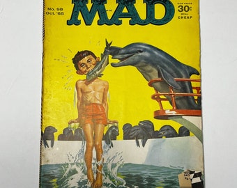 May include: A vintage MAD Magazine cover, issue No. 98 from Oct. '65, features a cartoon illustration. A boy in red shorts is in the water, a dolphin is on a boat, and the boy is being fed a fish. The cover is yellow with the magazine's logo.