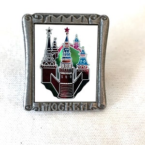 Puede incluir: Un pin rectangular de metal con una imagen detallada de un castillo con torres rojas, negras y azules, y un fondo verde. El pin tiene la palabra "MOCKBA" en la parte inferior y "TIMM" en la parte superior.
