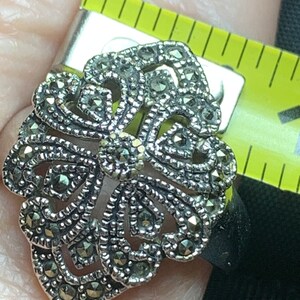 May include: A silver-toned marcasite ring with an ornate, openwork design. The ring features a central floral motif surrounded by swirling patterns and is accented with small, sparkling marcasite stones. The ring is being measured with a yellow measuring tape.