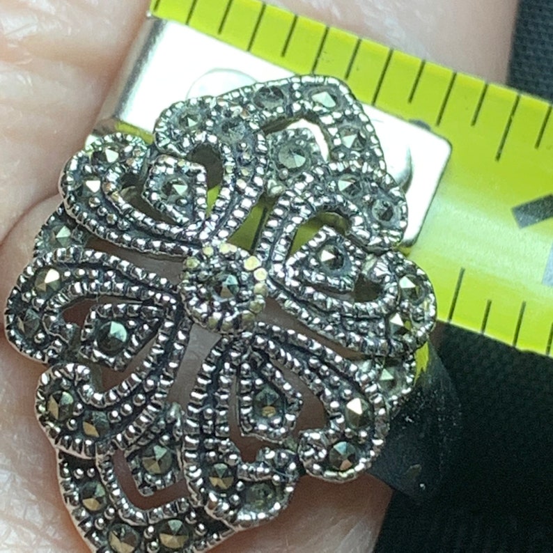 May include: A silver-toned marcasite ring with an ornate, openwork design. The ring features a central floral motif surrounded by swirling patterns and is accented with small, sparkling marcasite stones. The ring is being measured with a yellow measuring tape.