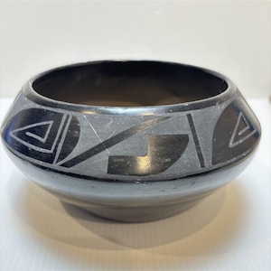 May include: A black ceramic bowl with a smooth, polished finish. The exterior features a wide band decorated with matte black geometric patterns, including triangles, spirals, and angular lines that wrap around the circumference of the vessel.