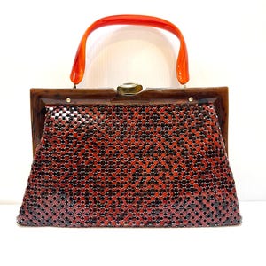 May include: A vintage handbag with a woven, reddish-brown body and a dark brown frame. The bag features a bright orange handle and a clasp closure. The overall design is reminiscent of mid-century fashion.