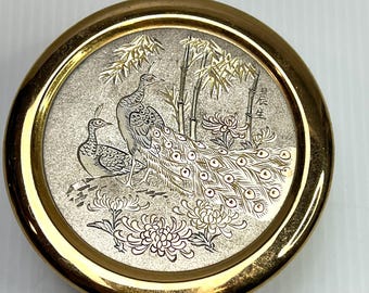 Könnte beinhalten: Ein rundes, goldfarbenes Metalldöschen mit einer strukturierten, silberfarbenen Einlage auf dem Deckel. Die Einlage zeigt zwei Pfaue, Bambusstangen und Chrysanthemenblüten sowie eingravierte chinesische Schriftzeichen auf der rechten Seite.