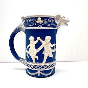 May include: A blue and white ceramic pitcher with a ram's head spout and a curved handle. The pitcher features white relief figures of cherubs and decorative patterns. The overall design is a classic, vintage style.