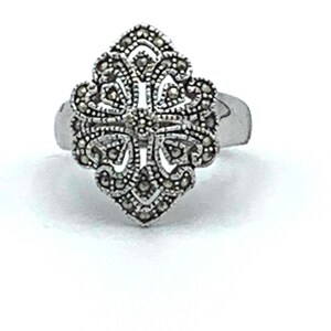 May include: A silver ring with a detailed, ornate design. The ring's centerpiece is a symmetrical, floral-like shape, encrusted with small, dark stones. The ring is set against a white background, highlighting its intricate details.