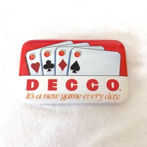 Puede incluir: Un pin rectangular rojo y blanco de DECCO con cartas de juego. El pin presenta cuatro cartas de juego con los palos de corazones, tréboles, diamantes y picas. El texto "DECCO" es rojo, y el lema "It's a new game every day" está en cursiva.