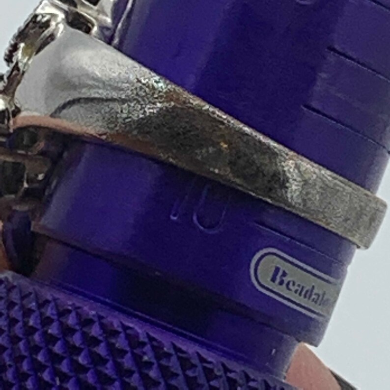 May include: Close-up of a silver ring with a textured band, resting on a purple ring sizer. The ring sizer has the number 10 visible. The ring has a decorative design on the top.