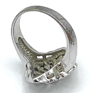 May include: Silver-toned ring with a textured band and a detailed, ornate design on the face. The ring's face features a complex pattern with small, raised elements, creating a vintage or antique aesthetic. The ring is set against a white background.