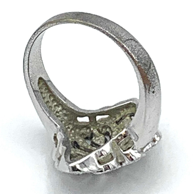 May include: Silver-toned ring with a textured band and a detailed, ornate design on the face. The ring's face features a complex pattern with small, raised elements, creating a vintage or antique aesthetic. The ring is set against a white background.