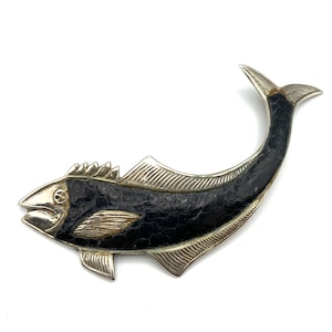 Puede incluir: Broche plateado con forma de pez y cuerpo negro texturizado. El broche presenta aletas detalladas, una cola y una cabeza con la boca abierta. El diseño es una representación estilizada de un pez, adecuado para añadir un toque único a cualquier atuendo.