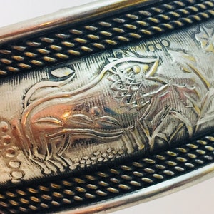 May include: Silver-coloured metal bangle bracelet with detailed engraved floral and animal designs. The bracelet features a pattern of leaves, flowers, and a stylized animal figure. The edges are accented with a textured, gold-coloured trim.