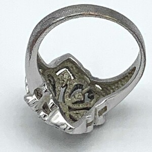 May include: Silver-toned ring with an ornate, openwork design. The ring features a detailed, textured pattern on the face and band. The ring is set against a white background, highlighting its intricate details. A piece of jewelry.