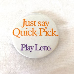Puede incluir: Un botón redondo blanco con el texto "Just say Quick Pick." en naranja y "Play Lotto." en morado. El botón es un artículo promocional, probablemente para una lotería o un servicio de apuestas.