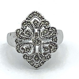 May include: A silver ring with an ornate, floral-inspired design. The ring's centerpiece features a detailed pattern of small, dark stones, creating a textured, eye-catching effect. The band is a simple, polished silver.