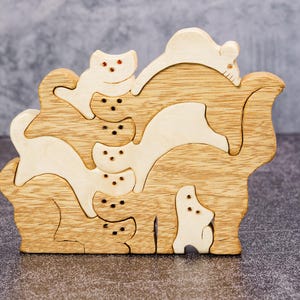 May include: A wooden puzzle featuring a stack of stylized cats in various poses. The puzzle is made of light and dark wood pieces, with the cats' eyes represented by small red dots. The overall design is a playful and decorative item.