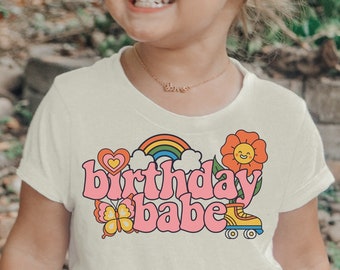 May include: A young child wearing a light yellow t-shirt with a rainbow, heart, flower, and roller skate graphic and the text "birthday babe". The child is also wearing pink shorts with white trim.