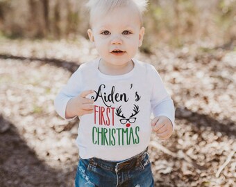 May include: A white long-sleeved bodysuit with the text "Aiden's First Christmas" in black and green lettering. A red-nosed reindeer with glasses is depicted above the text.