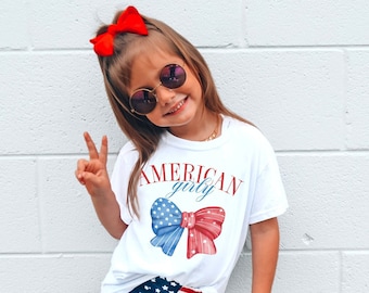 May include: A white t-shirt with a red, white, and blue bow and the text "AMERICAN girly" printed on it. The shirt is being worn by a child wearing red, white, and blue star-patterned shorts and sunglasses.