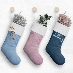 Puede incluir: Tres calcetines de Navidad colgados en una pared blanca. Los calcetines son azules, rosas y azul marino. Cada calcetín tiene un nombre: "Abigail", "Ashley" y "Ashley".