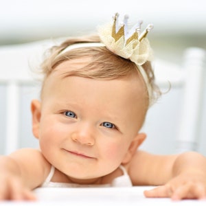 May include: A baby girl with blonde hair wearing a white headband with a gold crown and tulle. The baby is smiling and looking at the camera.