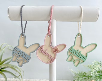 May include: Three wooden bunny-shaped Easter ornaments with personalized names, Grayson, Aubrey, and Beckham. Each ornament is painted in a different color: blue, pink, and green. The ornaments are hanging from a white stand.