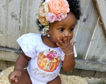 May include: A baby girl wearing a white onesie with a pumpkin and the text "I am one" in gold glitter. She is wearing a flower crown with peach, pink, and cream flowers. She is sitting on a bale of hay.